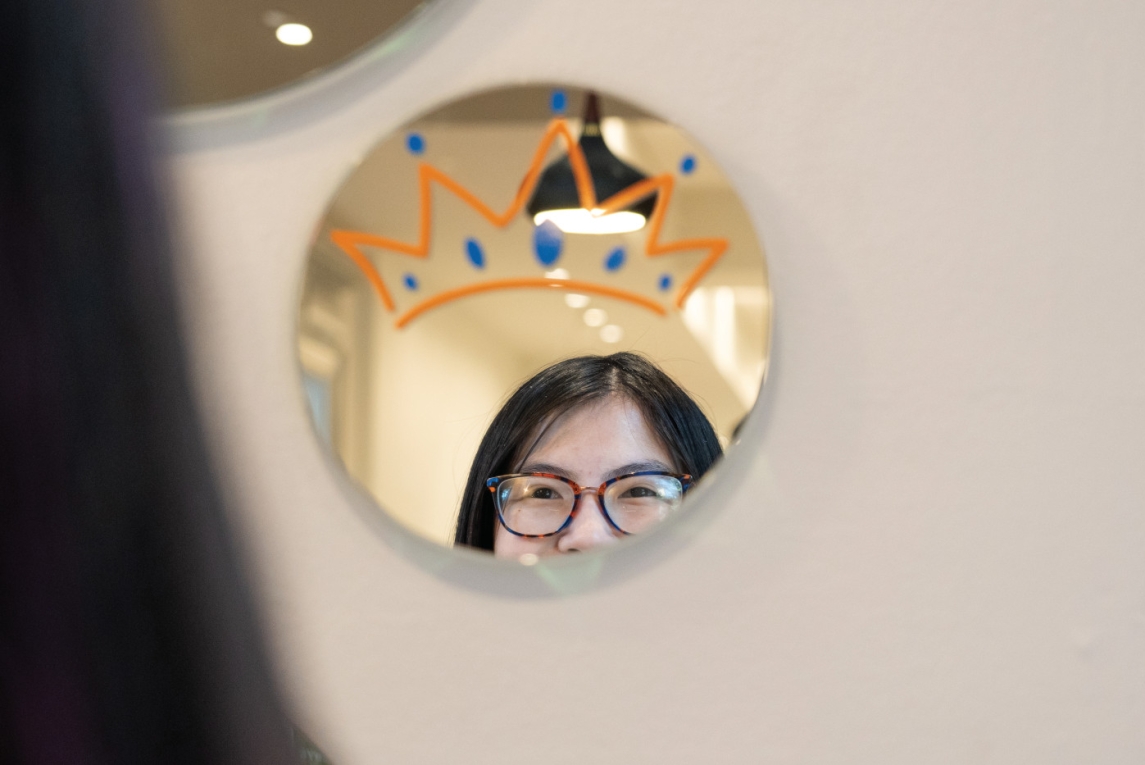 the upper part of a person's face is visible reflecting in a circular mirror with a crown painted on it