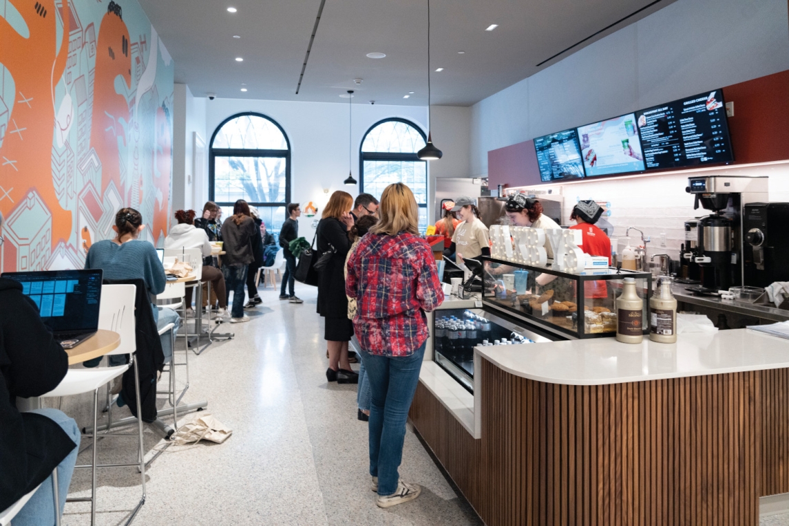 Saxby's cafe interior at the UArts student center. a coffee bar at the right with busy baristas behind it is bustling as people line up to order from menu screens hanging above and people sit with laptops at the left mural-painted wall