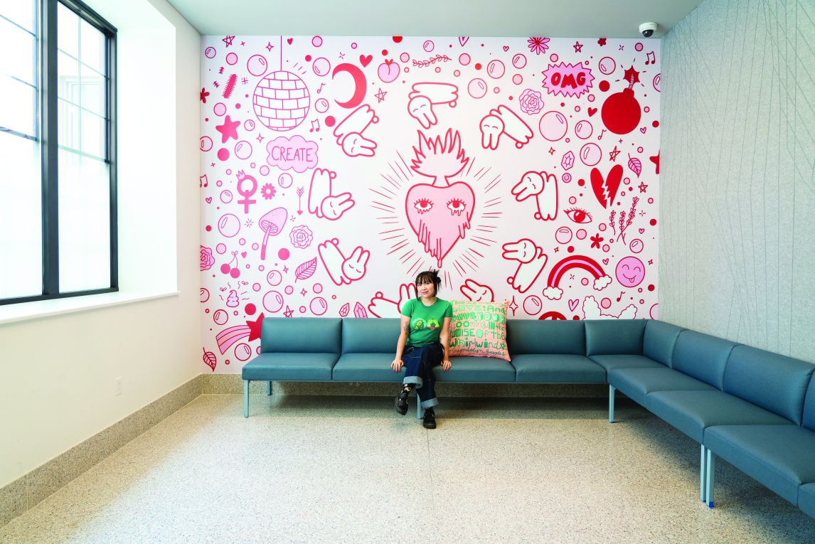 interior of the uarts student center, with a person i na green shirt seated on a green wrap-around series of couches ringing a room against a white wall decorated with pink and red line drawings of cute and bubbly imagery, focused on a heart with eyes