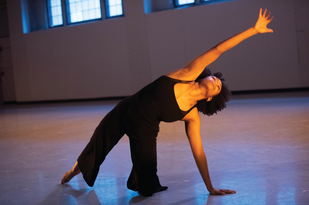 a person in a black outfit of a tank top and loose pants strikes a dynamic stretching pose on a blank gray dancefloor in a warm spotlight