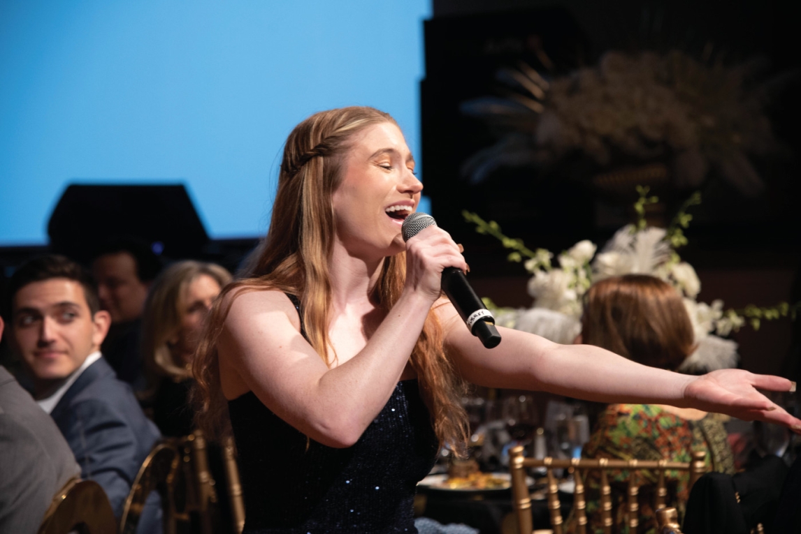 a person with long blond hair in a black dress is singing into a handheld microphone in a busy seated setting with decorated tables and gold-colored chairs surrounding