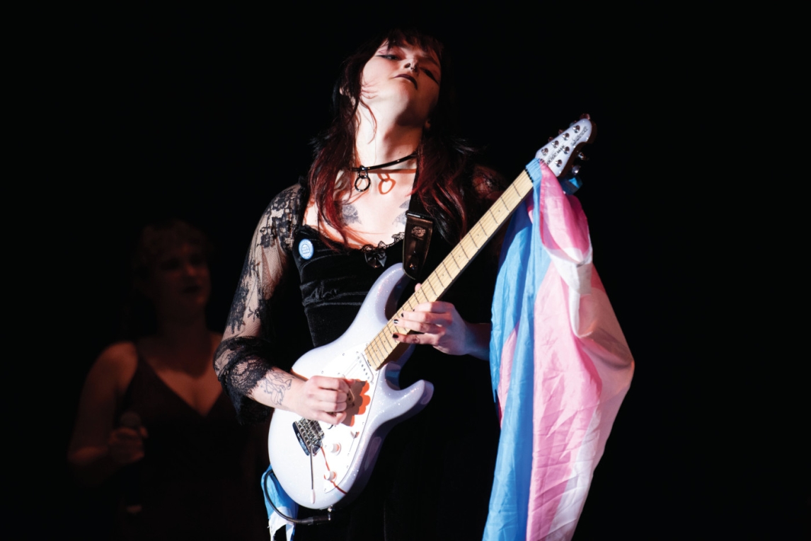 a person with a white guitar with a trans pride flag hanging from it plays in a stark spotlight
