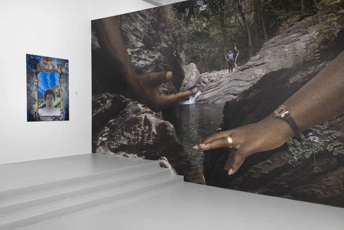 Installation view of a large vinyl mural depicting two Black hands reaching towards one another, displayed next to a vinyl printed portrait of a Black woman