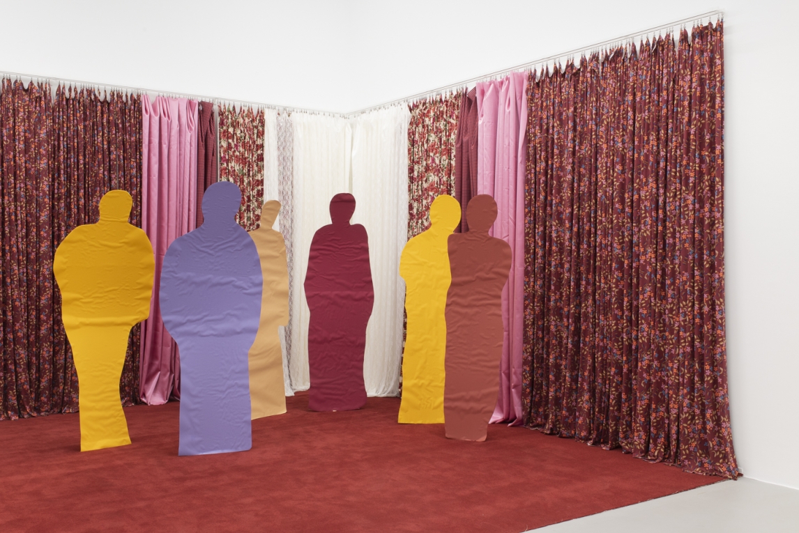 Installation view of gallery with various curtains in the background and human-shaped cut-outs covered in bright paper in the foreground
