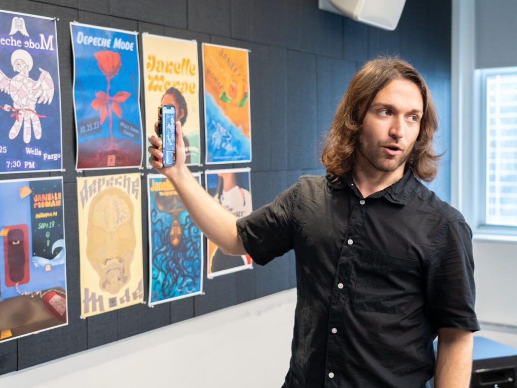a person in a black button up with long auburn hair and a scruffy beard holds a phone up to a wall of posters while looking back away from the wall at someone out of the frame. 