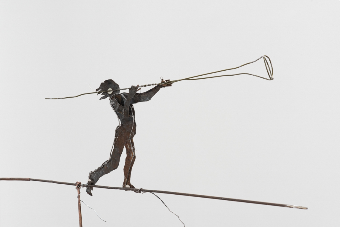 Sculpture of metal figure holding wire horn while standing on wire balanced on metal wire base