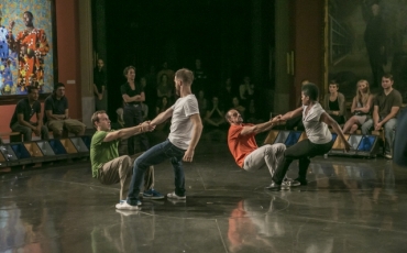 Four men dancing, in which two men are pulling up their partners while audience members sit around them. 