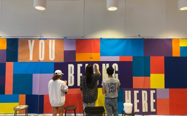 Three students working on a fabric mural made of multicolored squares and rectangles with the words You Belong Here in large white type on blocks of orange and purple color