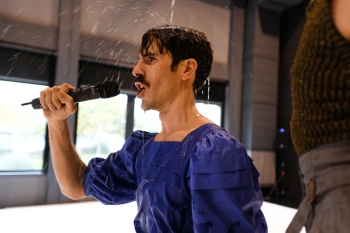 Albert Quesada holds a microphone and performs as water falls on to him from above and out of the frame on