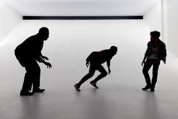 Three actors appear performing in silhouette on a white stage set