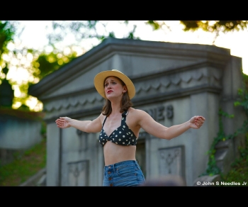 Julia Kershetsky performing outdoors with her arms outstretched in a blue and white dotted halter top and a straw hat