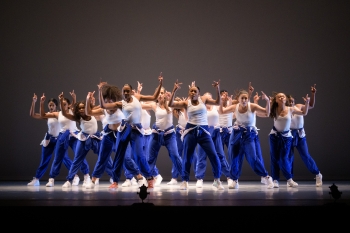 A group of dancers stand on a stark stage dressed in bright blue pants and matching white tanktops and they all have their arms raised