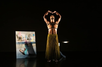 Uwazi Zamani performing on a dark stage and wearing a leather harnesslike chest piece and a pleated long skirt in mustard color