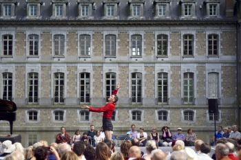Albert Quesada performs on an outdoor stage and has one arm raided and one stretched out in front of while wearing a red shirt and black shorts