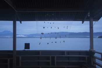 A light netting with small abstract objects attached to it that is hung on a structure overlooking the water 