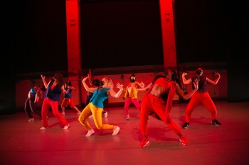A group of dancers lit with bright red lights bend their knees and backs backward and hold their arms up 