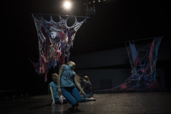 Three dancers in various poses on a stage where two are kneeling and one appears to be falling in front of a multicolored fabric backdrop that resembles spiderwebs
