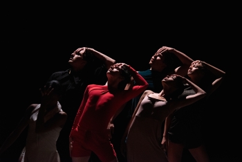 Five dancers on a dimly lit stage look up and over their shoulders and their hands are placed on their heads