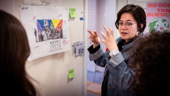 Rina Alfonso teaching a class in front of a whiteboard that displays a layout design and sketch of human silhouettes with the words Strike No Mail No Pay No Benefits at the bottom 