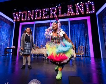 An actor dressed in a school uniform and another in a multicolor tulle skirt and bright green sneakers and a light purple wig skirt standing on a stage lit with blue and purple that has the word Wonderland in lights above a table and chairs 