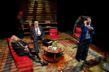 A stage set of a living room with a red couch and two different kinds of read chairs and two actors sitting while one is standing and talking on the phone