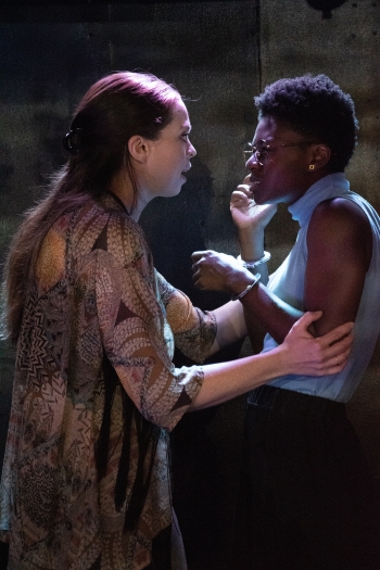 Two actors looking intently at one another while obne is wearing a loose printed dress and the other is wearing a light blue sleeveless turtleneck