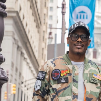 A student veteran dressed a camouflage jacket with patches stands on Broad Street in front of a blue and white UArts banner