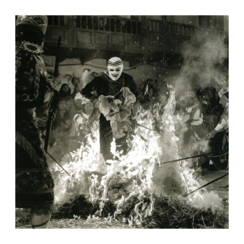 A black and white image that includes a subject in a white mask with black stripes walking through a fire on the street