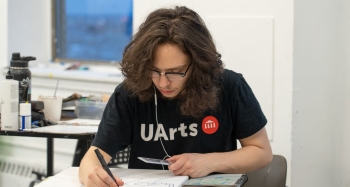 A painting student works at a desk while wearinng a UArts tee shirt and using headphones