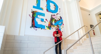 A student designer dressed in a red shirt and black pants standing in front of their mural in the Student Center 
