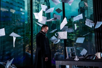 An actor dressed in a suit on a stage set like a high rise office building with papers flying around them