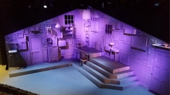 A stage set of the interior of a house lit entirely in purple with a floor lit in light blue