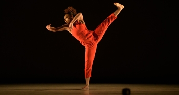 A dancer dressed in an orange jumpsuit extends a leg high in the air with their foot flexed