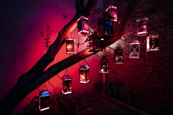 A work by Dilmar Gamero that includes lanterns with images on them and all the lanterns hanging on a tree branch in a room lit with red and featuring a brick wall