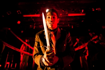An actor dressed in a suit and lit with red light holds a dagger 