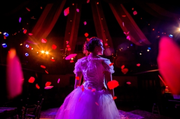 A performer stands onstage lit with red and purple lights while confetti falls around them
