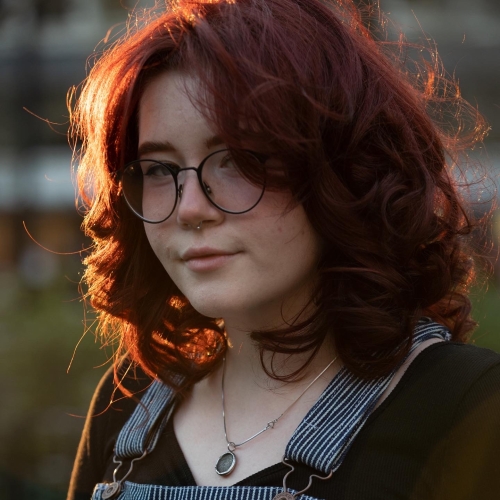 portrait of Aarin Wilson. She has shoulder0length red hair, black round glasses, a small septum ring and is wearing striped overalls. sunlight is illuminating her hair from the left. 
