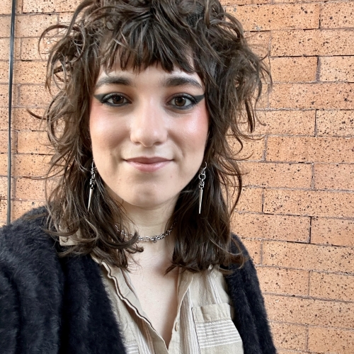 headshot of gabby bogdanoff with a background of faded bricks. Gaby is wearing a fuzzy black cardigan, and has frizzy hair and sharp eyeliner wings and pointed chain earrings. 