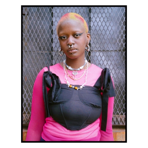 portrait of Zeinab Diomande, wearing a pink long-sleed shirt with a black corset-like garment, standing against a metal mesh window. Zeinab has several large gauge earrings and septum piercing and her buzzed hair is dyed yellow and red