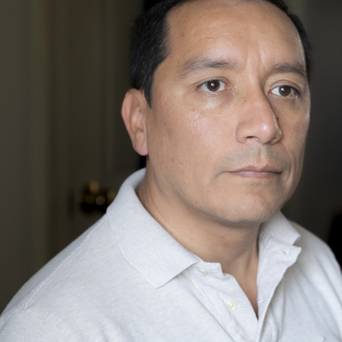 A closeup of Dilmar Gamero looking away past the camera and wearing a white collared shirt in front of a dark background 