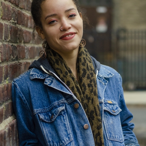 Noa Denmon in a blue denim jacket and a leopard print scarf leaning against a brick wall and smiling