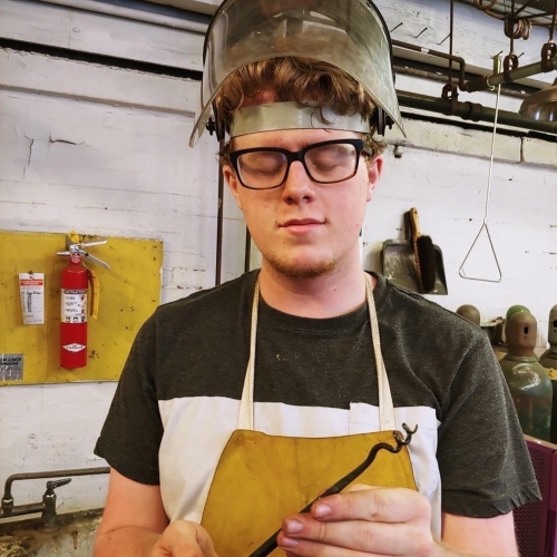 Portrait of Logan Smith in protective gear while working on sculpture. Smith is holding a corkscrew-like tool, is wearing a yellow apron, and has a visor on head