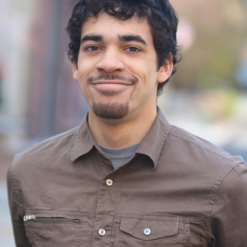 headshot of anthony martinez-briggs. Anthony is show smiling against a blurred out of focus outdoor background. Anthony has curly black hair, a chinstrap bears, and is wearing a brown button down shirt with double breasted pockets.