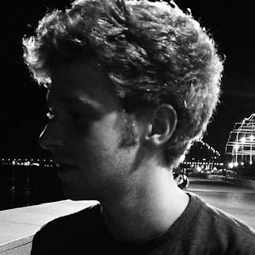 A grayscale photograph of Adam Samson BFA ’14 (Multimedia); he is lit from behind so his faceis obscured. It seems to be night and there are lights in the background.