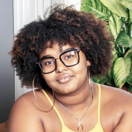 A medium-dark skinned young woman with thick, black-framed glasses and large hoop earrings wearing a silver necklace with what looks like a wishbone as a pendant is smiling slightly looking directly at the camera with a plant behind them.