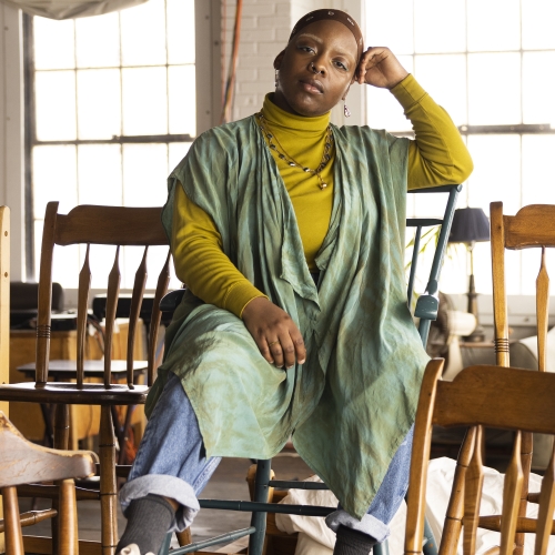 angel edwards sitting in a room with wooden chairs wearing a mustard turtleneck with a green smock over it and jeans 