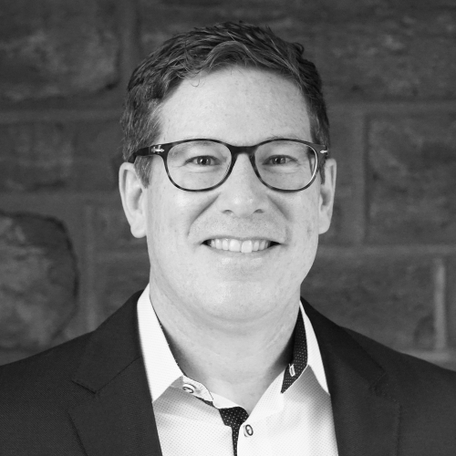 A grayscale portrait of Brian Yetzer, faculty in the Interaction Design program. Brian is smiling at the camera and wearing dark-framed glasses, a button-up shirt and a suit jacket.