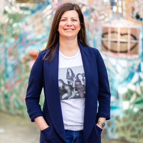 Jen Corsilli in a band tee shirt and a blue blazer and blue jeans standing in front of a mosaic made of mirrors and ceramic tiles