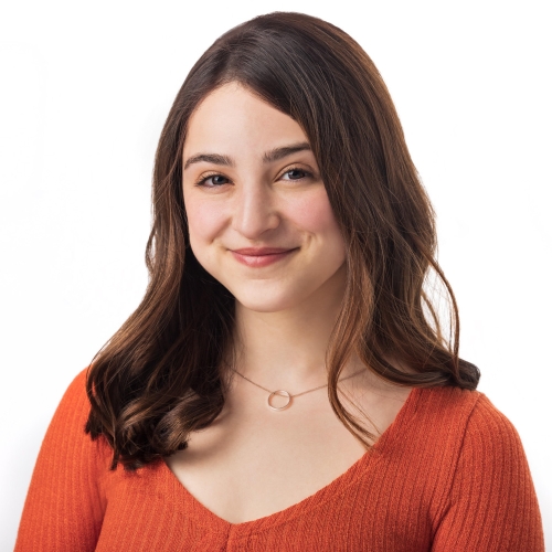 headshot of  Ella Kahan against a blank white background. Ella has long brown hair, is wearing a deep V orange sweater, has a golden ring on a subtle chain and is smiling warmly at the viewer