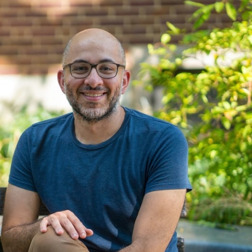 Dr. Abdelmagid wears glasses and a blue t-shirt while sitting outdoors on a park bench in the shade. Behind him are out of focus green shrubs.